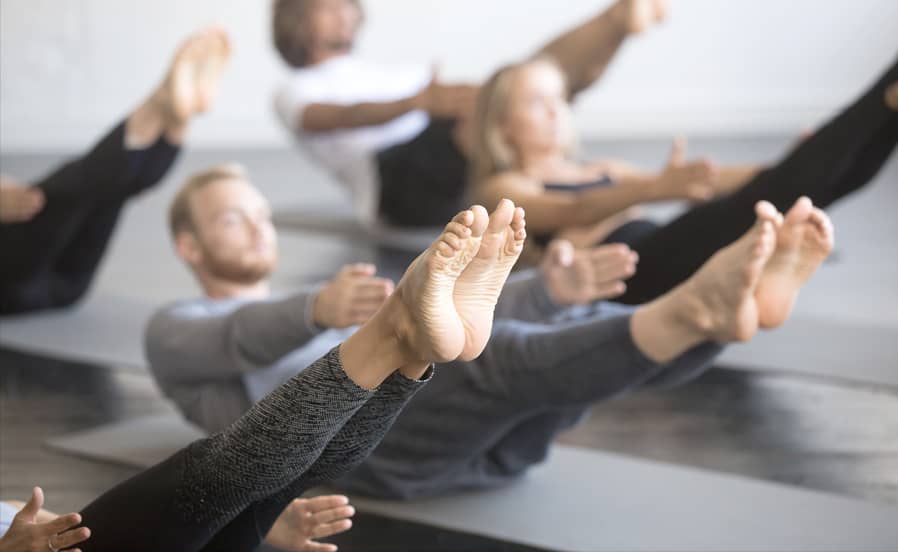Mehrere Personen halten eine kraftvolle Yoga-Balanceübung mit angehobenen Beinen auf Matten im Kursraum.