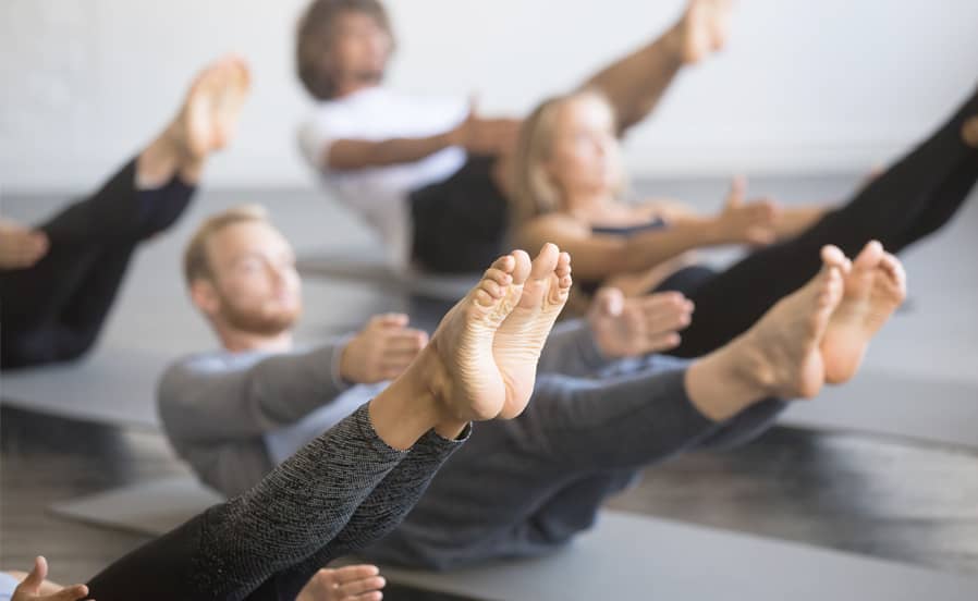Mehrere Personen halten eine kraftvolle Yoga-Balanceübung mit angehobenen Beinen auf Matten im Kursraum.