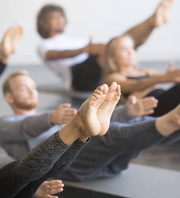 Mehrere Personen halten eine kraftvolle Yoga-Balanceübung mit angehobenen Beinen auf Matten im Kursraum.