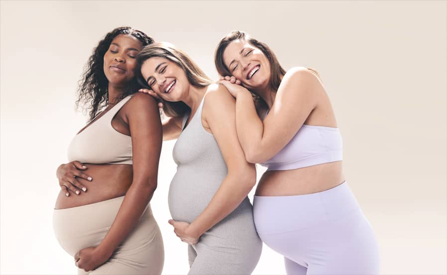 Drei schwangere Frauen lächeln, lehnen sich aneinander und halten liebevoll ihre Babybäuche.