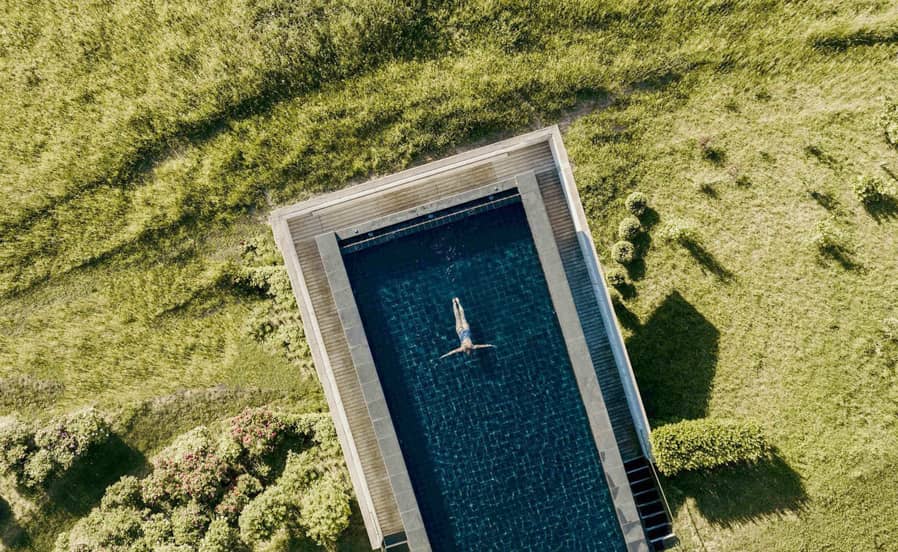 Luftaufnahme eines rechteckigen Outdoor-Pools inmitten einer grünen Wiese der Outdoor-Spa-Anlage des Naturhotels Pfösl; eine Person treibt entspannt auf dem Rücken im klaren Wasser.