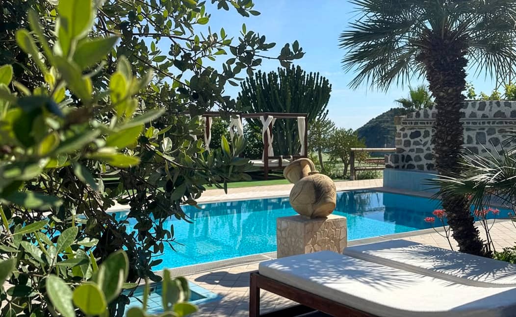 Sonniger mediterraner Poolbereich mit Liege, Skulptur und üppiger Vegetation; im Hintergrund sind Pergola, Steinmauer und Meerblick zu sehen.