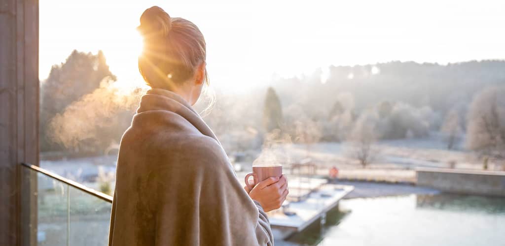 Frau in eine Decke gehüllt genießt bei Sonnenaufgang mit einer dampfenden Tasse Tee den Blick auf den winterlich-frostigen Pfälzer Wald.
