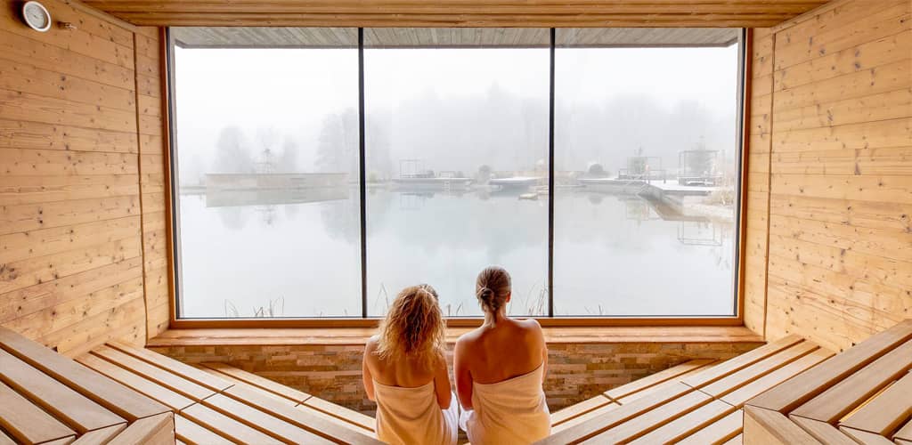 Zwei Frauen sitzen in der holzverkleideten Panoramasauna blicken durch das große Fenster auf den nebelverhangenen Naturbadeteich des Pfalzblick Wald Spa Resort.