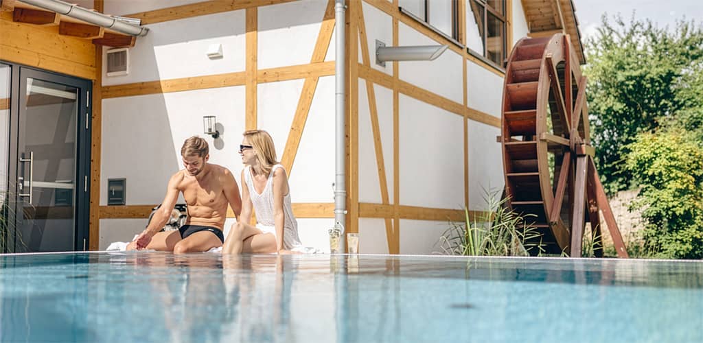 Paar sitzt entspannt am Außenpool vor Fachwerkgebäude mit großem Wasserrad und genießt die Sonne.