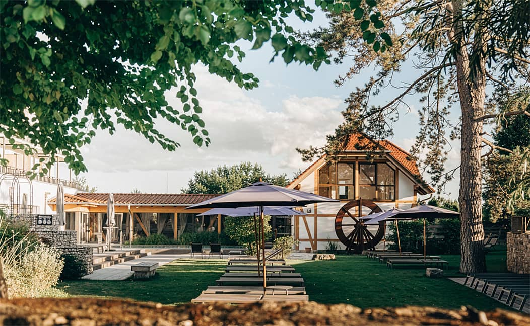Die ruhige Hotelanlage von Jordan's Untermühle mit Terrasse, Liegen und Sonnenschirmen vor Gebäuden mit Holzelementen und Gartenanlage.