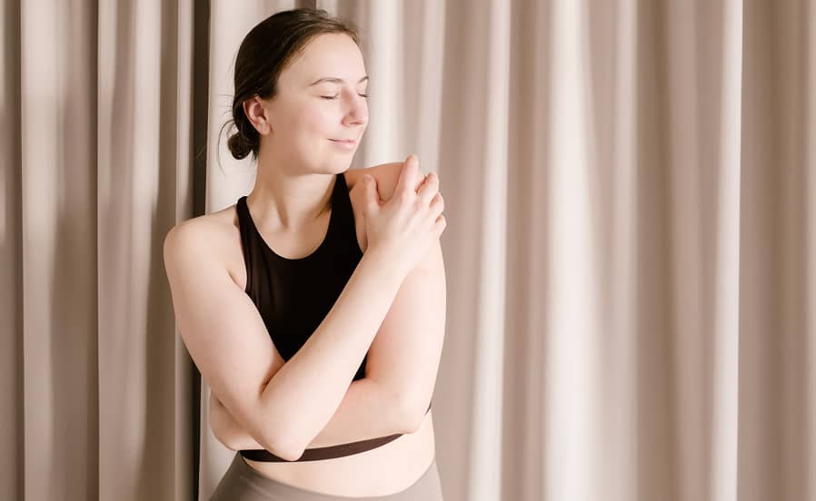 Yogalehrerin Yvonne Schatz steht in Sportkleidung mit geschlossenen Augen vor Vorhang und legt achtsam die Hand auf ihre Schulter.