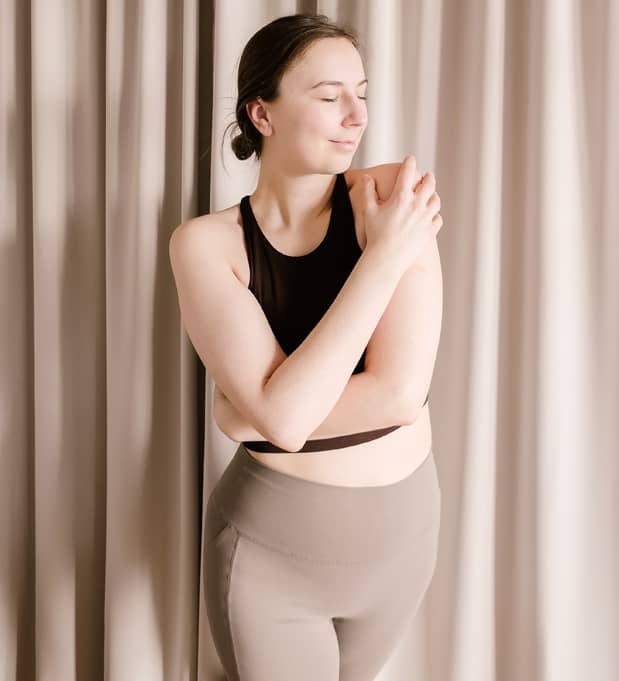 Yogalehrerin Yvonne Schatz steht in Sportkleidung mit geschlossenen Augen vor Vorhang und legt achtsam die Hand auf ihre Schulter.