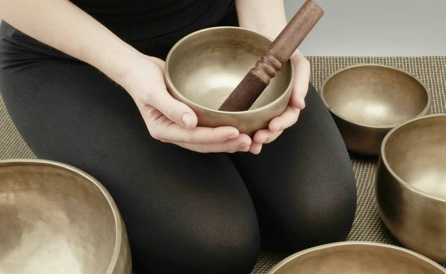 Eine Frau sitzt im Schneidersitz und hält eine Messing-Klangschale mit Holzklöppel, umgeben von weiteren Klangschalen auf einer Matte.