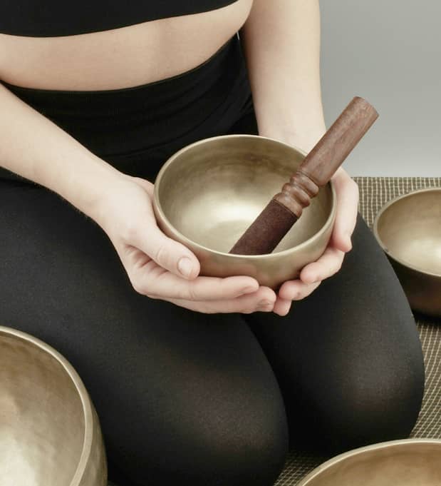 Eine Frau sitzt im Schneidersitz und hält eine Messing-Klangschale mit Holzklöppel, umgeben von weiteren Klangschalen auf einer Matte.