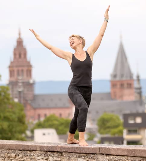 Yoga-Lehrerin Susanne Weisheit in dunkler Sportkleidung balanciert barfuß auf der Mauer der Mainzer Zitadelle und streckt freudig die Arme nach oben; im Hintergrund sind unscharf der Hohe Dom u Mainz und die Altstadt zu sehen.