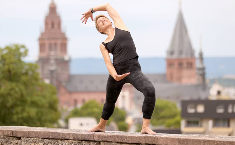 Yoga-Lehrerin Susanne Weisheit Frau in schwarzer Sportkleidung macht eine dynamische Yoga-Seitbeuge auf einer Mauer der Mainzer Zitadelle; im Hintergrund sind unscharf der Hohe Dom u Mainz und die Altstadt zu sehen.