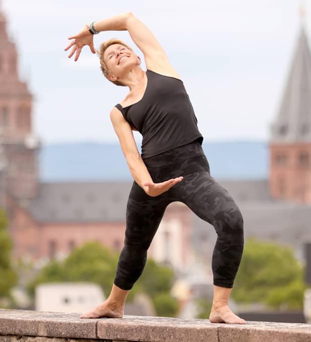 Yoga-Lehrerin Susanne Weisheit Frau in schwarzer Sportkleidung macht eine dynamische Yoga-Seitbeuge auf einer Mauer der Mainzer Zitadelle; im Hintergrund sind unscharf der Hohe Dom u Mainz und die Altstadt zu sehen.
