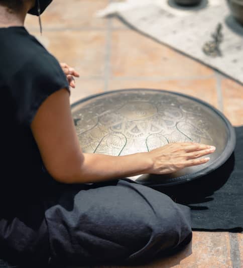 Person mit schwarzem ärmellosen Oberteil sitzt im Lotussitz auf einer dunkelgrauen Filzmatte und spielt auf einer vor seinem Schoß ruhenden mit Gravuren reich verzierten Handpan
