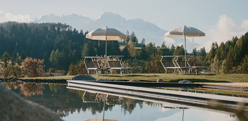 Liegestühle mit Sonnenschirmen am Naturpool des Naturhotels Pfösl in Deutschnofen, dahinter dichter Wald und ferne Berggipfel – ruhige Outdoor-Szene für Entspannung und Retreat.