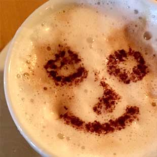 Bild zeigt eine große Tasse mit Kaffe von oben mit Milchschaum, auf dem ein mit Kakaopulver gezeichnetes Smiley lächelt (Foto: © Yogaplus/Susanne Weisheit)