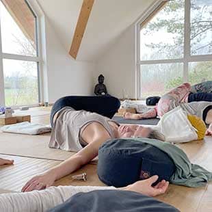 Die Teilnehmer des Yogaplus-Mini-Retreats im hellen Yogaraum von Jordan's Untermühle in seitlich liegender Drehung und mit ausgestreckten Armen auf dem Rücken und entspannen mit geschlossenen Augen auf ihren Yogamatten. (Foto: © Yogaplus/Susanne Weisheit)