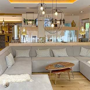Blick in die helle Spa Lounge von Jordan's Untermühle mit hellem Holzdielenboden, weiß verputzten Wänden über eine große hellgraue Loungesitzgruppe mit quergestreiften Kissen und modernen tief hängenden Drahtdeckenleuchtern auf die mit hellem Holz verkleidete Bar (Foto: © Yogaplus/Susanne Weisheit)