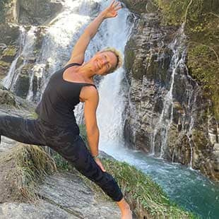 Yogalehrerin Susanne Weisheit in schwarzem Yogaoutfit praktiziert die Asana 'Krieger zwei' vor dem Bad Gasteiner Wasserfall. (Foto: © Yogaplus/Susanne Weisheit)