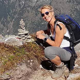 Yogalehrerin Susanne Weisheit sitzt im Tourenoutfit mit Rucksack und schweren Bergwanderschuhen neben einem kleinen Steinturm am Wegesrand und lacht sonnenbebrillt in die Kamera. (Foto: © Yogaplus/Susanne Weisheit)