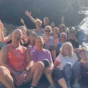 Gruppenbild der Teilnehmer:innen des Yogaplusretreats, wie sie zusammen auf einem großen Felsen vor den Bad Gasteiner Wasserfällen sitzen und in die Kamera grüßen. (Foto: © Yogaplus/Susanne Weisheit)