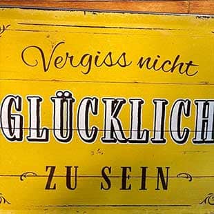 An eine Holzwand geschraubtes abgewetztes gelbes Blechschild mit der in unterschiedlichen Schrifttypen ausgeführtem Aufdruck '„Vergiß' nicht, glücklich zu sein'. (Foto: © Yogaplus/Susanne Weisheit)