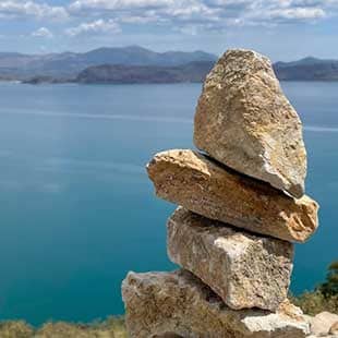Das unvermeidliche kleine Steintürmchen auf felsigem Boden bei sonnigem Wetter mit Blick auf die spiegelglatte See der Bucht von Kiveri (Foto: © Yogaplus/Susanne Weisheit)