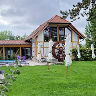 Grüne Liegewiese mit geschlossenen Sonnenschirmen vor einem Fachwerkhaus mit Wasserrad und angrenzendem Außenpool. (Foto: © Yogaplus/Susanne Weisheit)