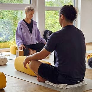 Teilnehmer eines Yogaretreats praktizieren gemeinsam Yoga in einem hellen Raum mit Holzboden, großen Fenstern und Blick ins Grüne. (Foto: © Yogaplus/Susanne Weisheit)