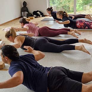 Yoga-Lehrerin Susanne Weisheit schult Retreat-Teilnehmer in einer Rückbeuge-Übung im hellen Yogaraum mit großen Fenstern, Holzfußboden und Gong im Hintergrund. (Foto: © Yogaplus/Susanne Weisheit)