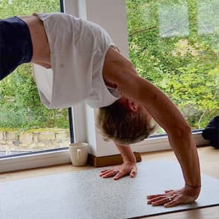 Yoga-Lehrerin Susanne Weisheit im hellen Yogaraum führt eine einbeinige Rückbeuge-Pose (Urdhva Dhanurasana) auf der Matte aus, mit Blick auf grüne Natur durch große Fenster. (Foto: © Yogaplus/Susanne Weisheit)