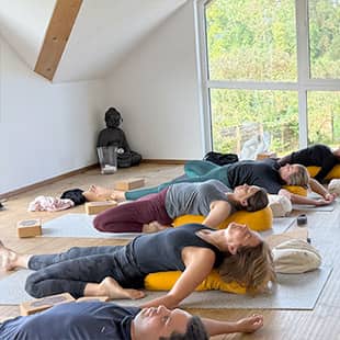 Retreat-Teilnehmer beim Yoga in einem hellen Raum, entspannt liegend auf gelben Bolstern, mit Blick auf grüne Natur durch große Fenster. (Foto: © Yogaplus/Susanne Weisheit)