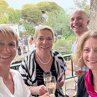 Vier lächelnde Personen stoßen auf einem Balkon mit Sektgläsern an, im Hintergrund eine grüne Gartenanlage mit weiteren Menschen. (Foto: © Yogaplus/Susanne Weisheit)
