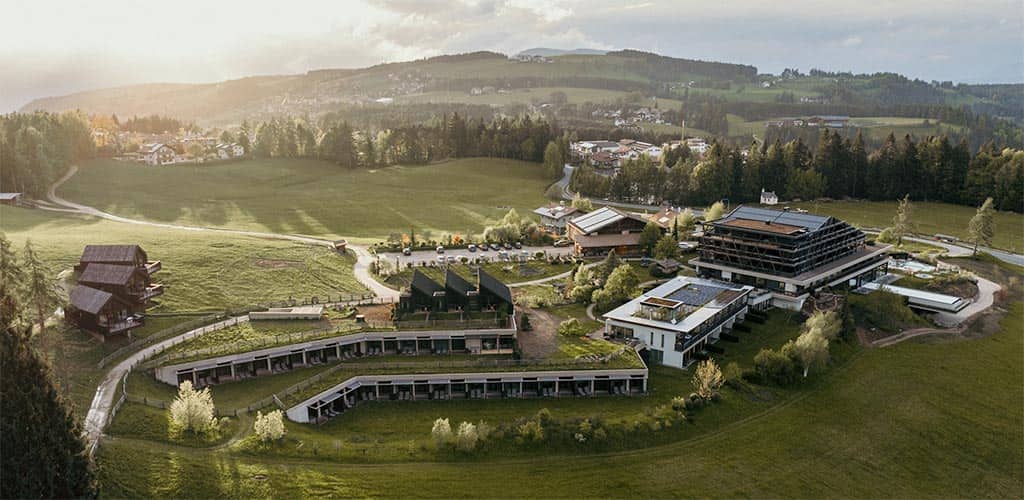 Luftaufnahme des weitläufigen Alpenresorts 'Naturhotel Pfösl' in Deutschnofen mit modernen Hotelgebäuden und Chalets, umgeben von grünen Wiesen, Wäldern und Hügeln im Abendlicht.