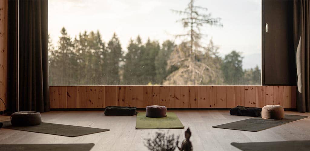Ruhiger Yogaraum im Naturhotel Pfösl mit großen Panoramafenstern zum Wald, auf dem Holzboden liegen Matten, Meditationskissen und gefaltete Decken.