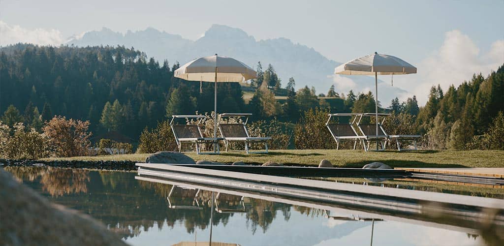 Liegestühle mit Sonnenschirmen am Naturpool des Naturhotels Pfösl in Deutschnofen, dahinter dichter Wald und ferne Berggipfel – ruhige Outdoor-Szene für Entspannung und Retreat.