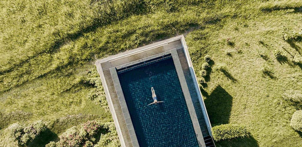 Luftaufnahme eines rechteckigen Outdoor-Pools inmitten einer grünen Wiese der Outdoor-Spa-Anlage des Naturhotels Pfösl; eine Person treibt entspannt auf dem Rücken im klaren Wasser.