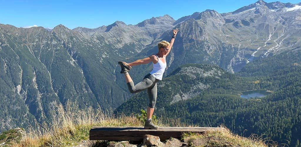 Yogalehrerin Susanne Weisheit in der Yoga-Pose Natarajasana auf einem Bergkamm, mit Wanderschuhen vor weitem Alpenpanorama und Tal mit Bergsee unter blauem Himmel.