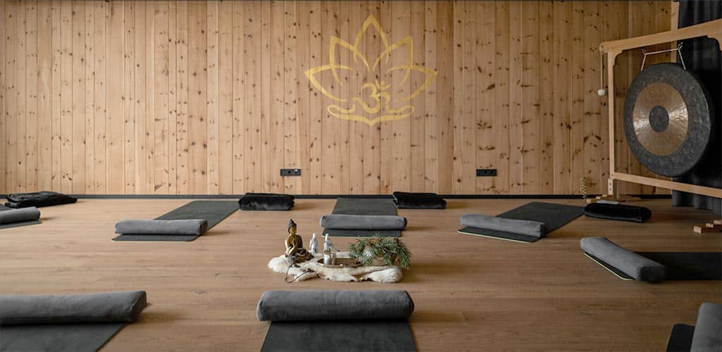 Ruhiger Yoga-Raum im Naturhotel Pfösl mit Holzvertäfelung, ausgerollten Matten und Bolstern, kleinem Altar mit Buddhafigur in der Mitte und großem Gong rechts; goldenes Om-Lotus-Symbol an der Wand.