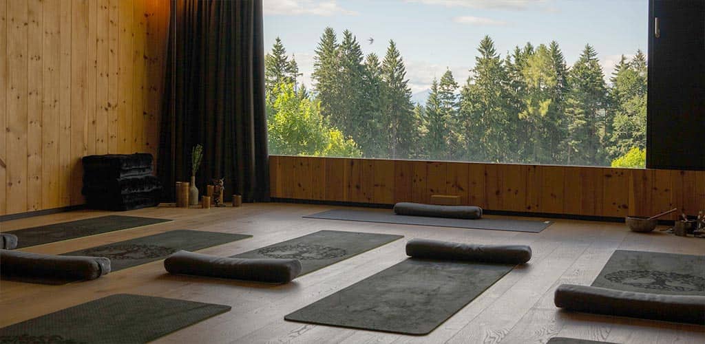 Yoga- und Meditationsraum im Naturhotel Pfösl mit Yogamatten und Sitzkissen, großem Panoramafenster mit Blick auf Waldlandschaft.