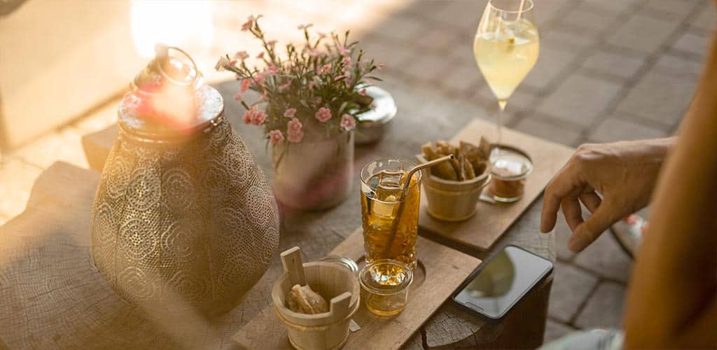 Nahaufnahme eines gedeckten Outdoor-Tisches mit Eistee im Glas, kleinen Snacks in Schälchen, dekorativer Laterne und Blumen; eine Hand greift nach dem Smartphone.
