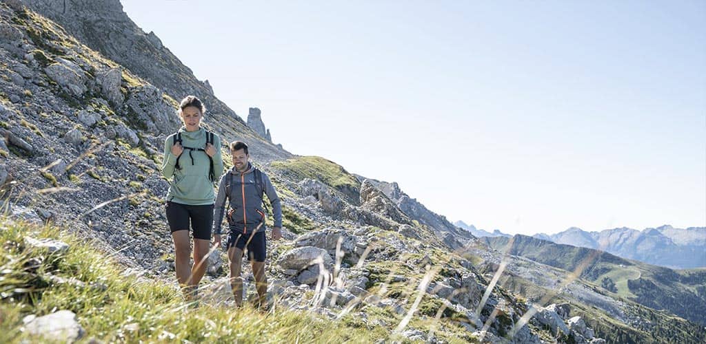 Zwei Wandernde mit Rucksäcken auf felsigem Alpenpfad im Rosengarten, umgeben von grünen Hängen und weitem Bergpanorama unter blauem Himmel.