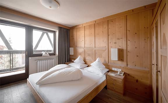 Gemütliches Doppelzimmer 'Old Style' im Naturhotel Pfösl mit Doppelbett, Holzvertäfelung an den Wänden, Nachttischen und Zugang zu einem Balkon mit Tageslicht.