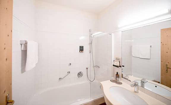 Das helle Badezimmer des Zimmertyps 'Old Style' im Naturhotel Pfösl mit Badewanne und Duscharmatur, großem Spiegel, Waschbecken und weißen Fliesen.