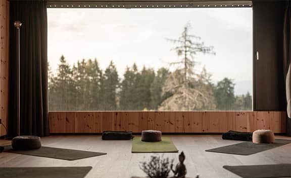 Yoga- und Meditationsraum im Naturhotel Pfösl mit Yogamatten und Sitzkissen, großem Panoramafenster mit Blick auf Waldlandschaft.
