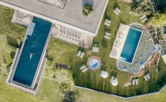 Luftaufnahme auf Wellnessbereich ‘Naturaspa‘ des Naturhotel Pfösl mit Infinity-Pool, Naturpool, Whirlpool und Liegewiese inmitten grüner Landschaft.