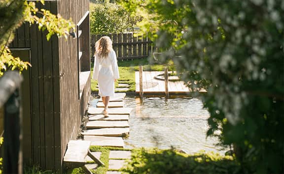 Frau im Bademantel geht auf Steinweg entlang eines Naturpools durch grüne Gartenlandschaft im Naturaspa des Naturhotels Pfösl.
