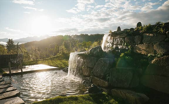 Natürlicher Gartenwasserfall mit Steinen und kleinem Teich im Sonnenlicht im 'Naturaspa & Pfösl Regeneration 360°', umgeben von grüner Landschaft und Fernblick auf bewaldete Hügel.
