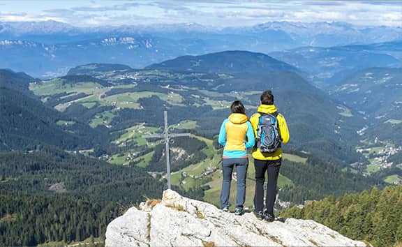 Zwei Wanderer mit bunten Jacken stehen auf einem Felsvorsprung neben einem Gipfelkreuz und blicken über das weitläufiges, grüne Eggental in Südtirol mit Wäldern und Dörfern.