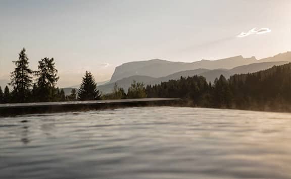 Blick über den Infinity-Pool im Naturaspa des Naturhotels Pfösl bei Sonnenuntergang, mit bewaldeten Hügeln und markanter Bergsilhouette im Hintergrund.